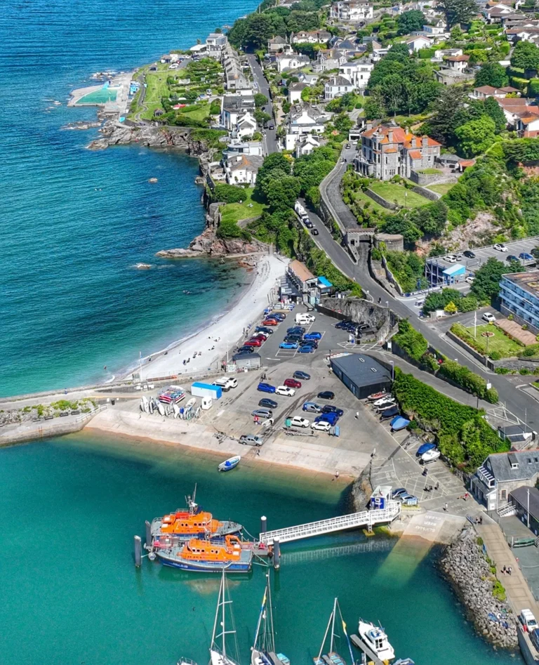 A scenic view of a sandy beach near Brixham, with gentle waves lapping the shore. In the background, Brixham Harbour is just a stone's throw away, showcasing colourful fishing boats and historic waterfront buildings under a bright blue sky.